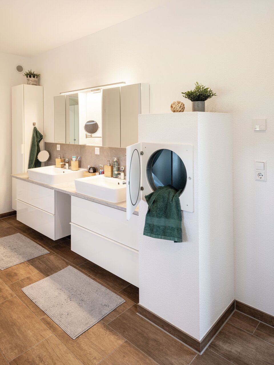 Modernes Badezimmer mit Doppelwaschtisch, großem Spiegel und integrierter Wäscheschachtlösung. Helle Wandfarben, Holzdetails und grüne Handtücher ergänzen das frische Design.
