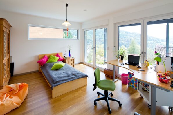 Modernes Kinderzimmer in einem frei geplanten FingerHaus mit Einzelbett, farbigen Kissen, großem Schreibtisch mit Stuhl und bodentiefen Fenstern mit Blick ins Grüne.