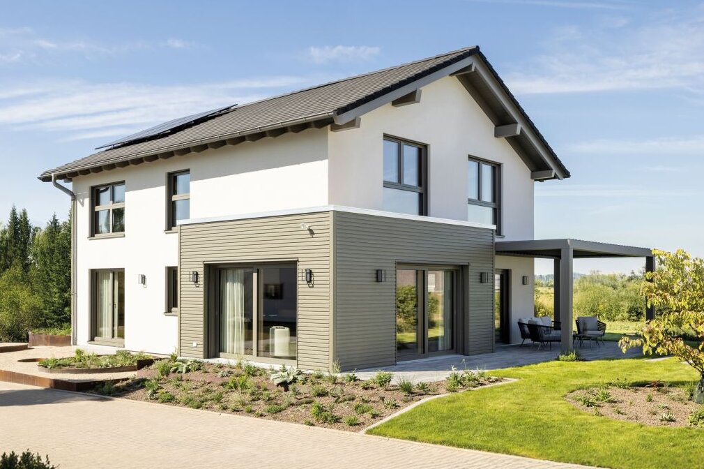 Zweigeschossiges modernes Haus mit Satteldach, weißer Fassade und grauen Holzverkleidungselementen, umgeben von bepflanztem Garten und gepflasterter Einfahrt, bei sonnigem Wetter fotografiert.