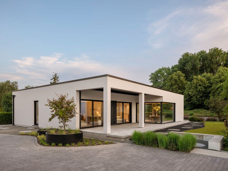 Musterhaus NIVO in Frankenberg mit moderner Bungalowarchitektur, weißer Putzfassade, überdachter Terrasse und großzügigen Eckfenstern in der Abenddämmerung.