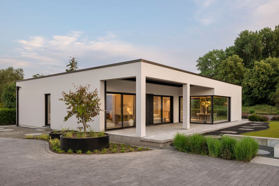 Musterhaus NIVO in Frankenberg mit moderner Bungalowarchitektur, weißer Putzfassade, überdachter Terrasse und großzügigen Eckfenstern in der Abenddämmerung.