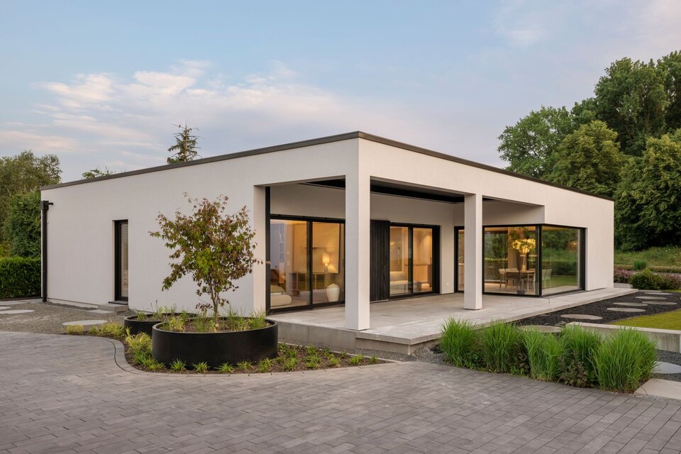 Musterhaus NIVO in Frankenberg mit moderner Bungalowarchitektur, weißer Putzfassade, überdachter Terrasse und großzügigen Eckfenstern in der Abenddämmerung.