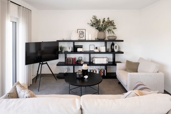 Wohnzimmer im Musterhaus TALO Fulda mit heller Einrichtung, modernem Regal, gemütlichen Sofas und großem Fernseher.