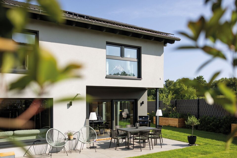 Doppelhaushälfte mit Satteldach, Terrasse mit Esstisch und Stühlen sowie Sitzmöbeln vor großen Terrassenfenstern