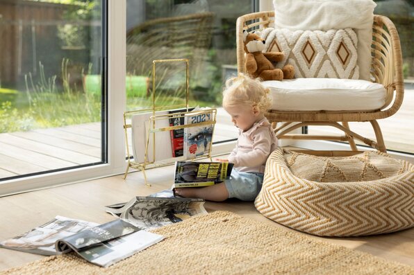 Gemütliche Leseecke in einem frei geplanten FingerHaus mit großem Fenster, hellen Holzböden und natürlichen Materialien. Ein Kind blättert entspannt in Magazinen und genießt das Sonnenlicht.