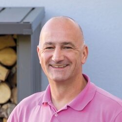 Ein Mann steht lächelnd an seinem Grill auf der Terrasse, im Hintergrund ist ein Holzlager und ein Gartenhaus zu sehen.