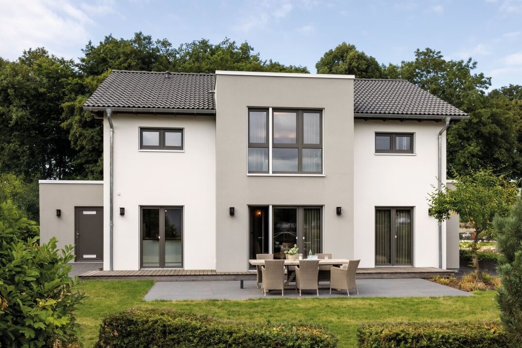 Zweigeschossiges Wohnhaus mit heller Putzfassade, symmetrischem Satteldach, mittigem Fassadenvorsprung mit großen Fenstern, Terrasse mit Tisch und Stühlen sowie Rasenfläche im Garten.