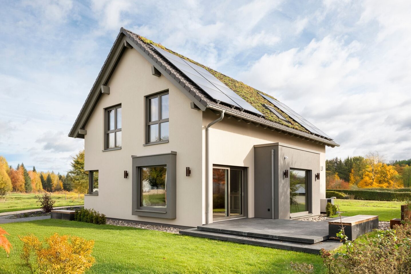 Zweigeschossiges Wohnhaus mit heller Putzfassade, Satteldach mit Photovoltaikmodulen und teilweise begrüntem Dachbereich, großen Fenstern, Terrassenbereich und Garten mit Rasenfläche.
