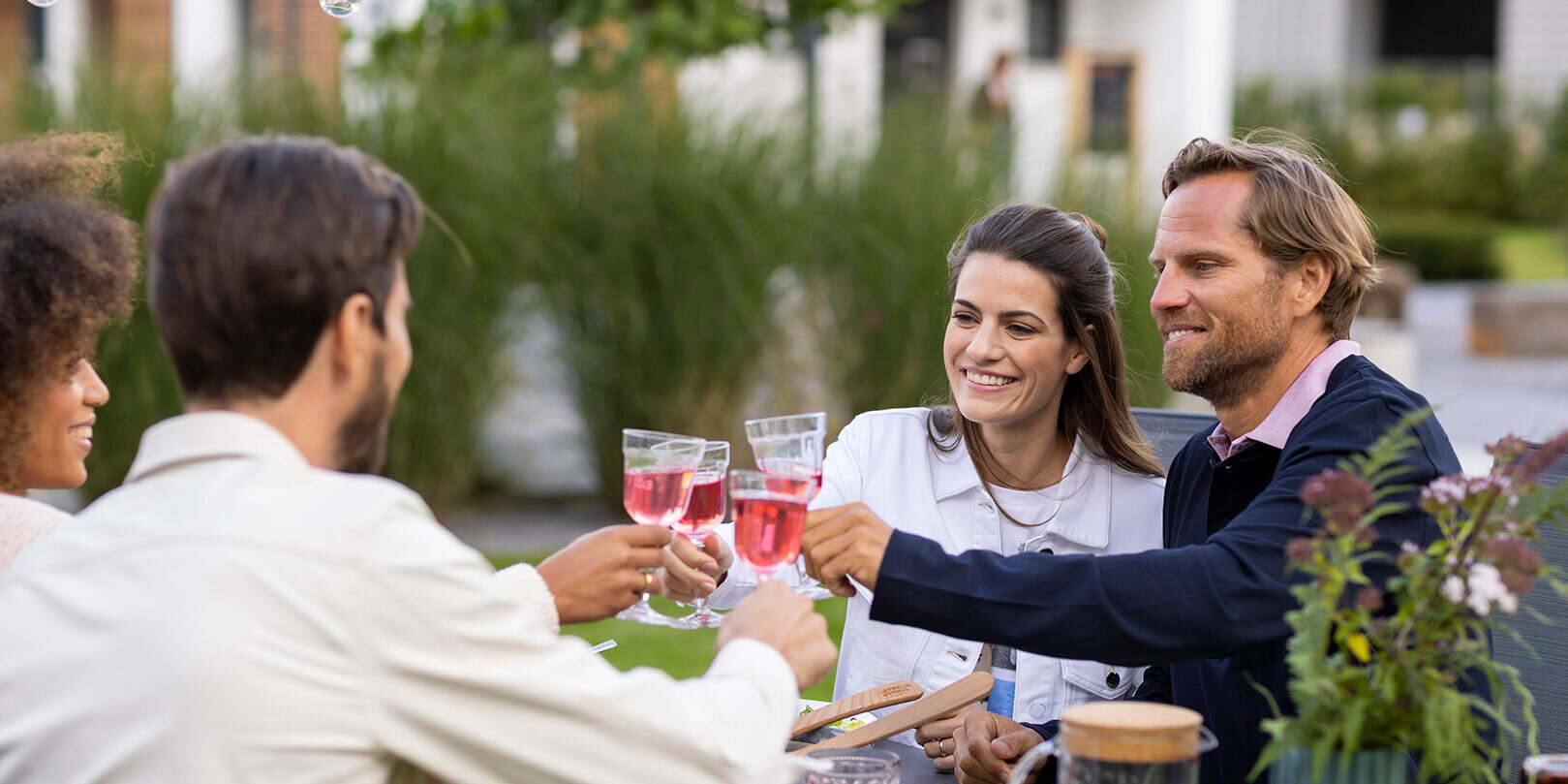 Vier Personen sitzen im Garten an einem Esstisch und stoßen mit Gläsern an. Auf dem Tisch stehen Speisen, Getränke und eine Vase mit Blumen. Die Stimmung ist fröhlich und entspannt.
