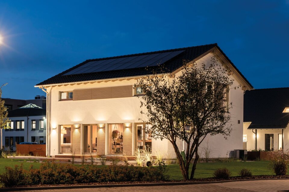 Abendliche Außenansicht des FingerHaus-Musterhauses SENTO 500 in Frankenberg mit weißer Putzfassade, dunkel gedecktem Satteldach und Photovoltaikmodulen; beleuchtete Fenster und Außenleuchten, davor Rasenflächen, Gehwege, Bäume und Straßenlaternen bei tiefblauem Nachthimmel.