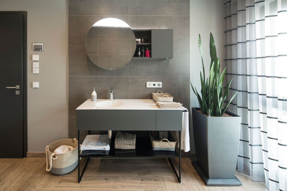 Modernes Badezimmer im Musterhaus ALTERO in Gießen mit Waschtisch, rundem Spiegel, dekorativer Pflanze und stilvoller Einrichtung.