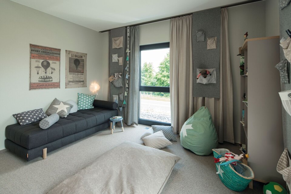 Gemütliches Wohnzimmer mit grauem Sofa, Kissen, Sitzsack, Vorhängen und dekorativen Wandbildern neben einer großen Fensterfront