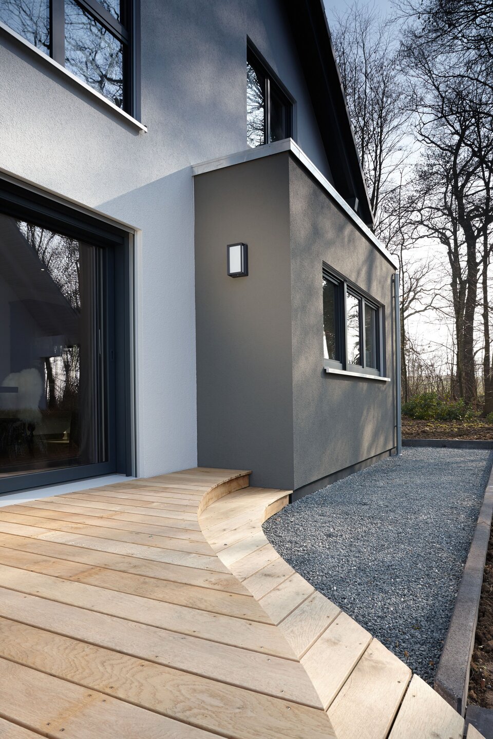 Detailaufnahme eines modernen Musterhauses mit grauer Fassade, großem Fenster und einer geschwungenen Holzterrasse. Daneben verläuft ein Kiesstreifen, im Hintergrund sind Bäume zu sehen.
