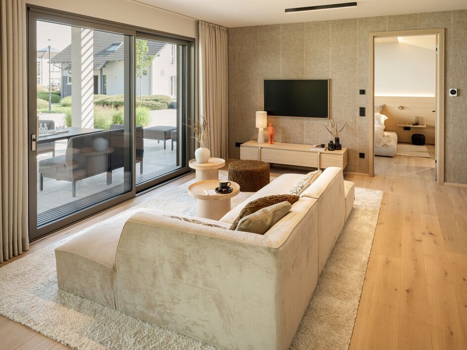 Wohnbereich im FingerHaus Musterhaus NIVO in Frankenberg mit hellem Ecksofa, großer Glasfront zur Terrasse und Blick ins Schlafzimmer.