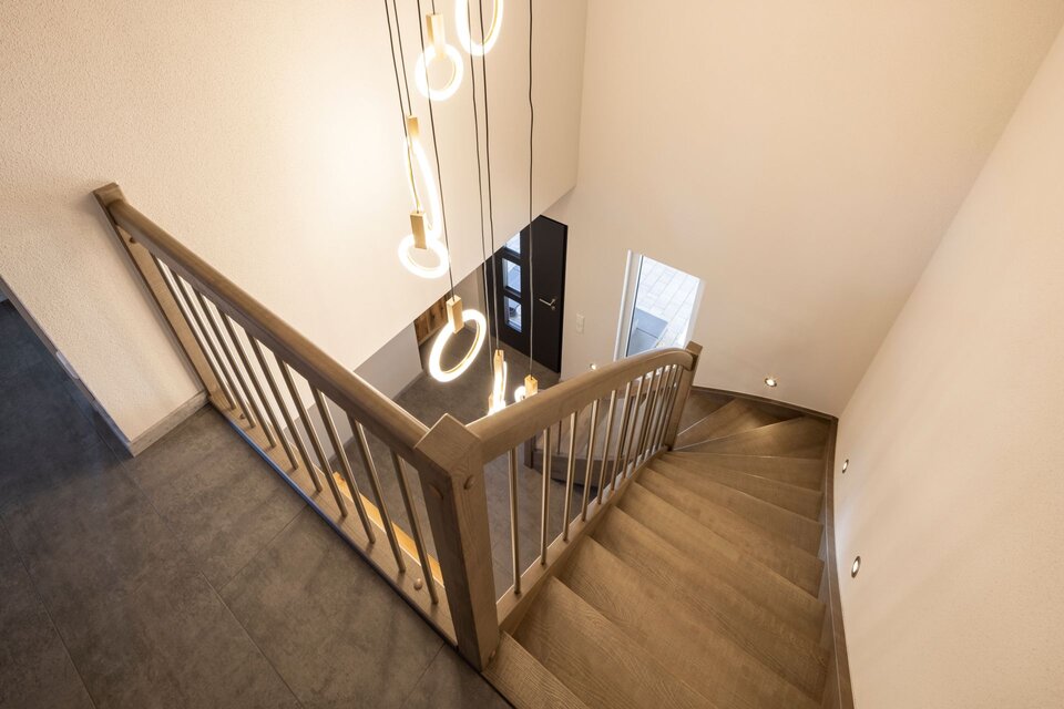 Blick von oben auf eine Holztreppe mit Metallgeländer und moderner Pendelleuchte. Hohe Decke, helle Wände und dunkler Boden im Eingangsbereich.