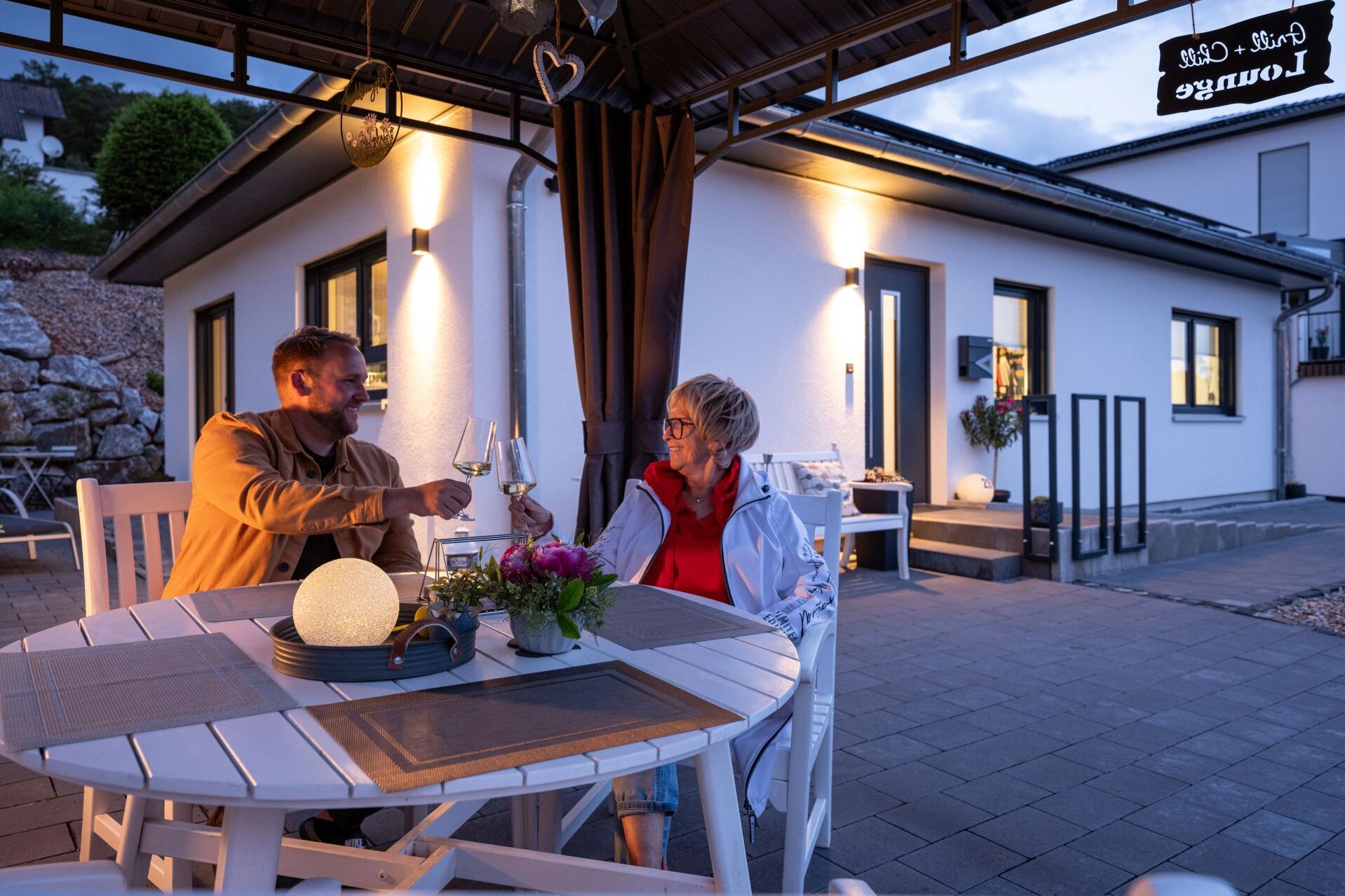 Zwei Personen sitzen am Abend in einem Pavillon vor einem Kundenhaus NIVO 80 und stoßen mit Weingläsern an.