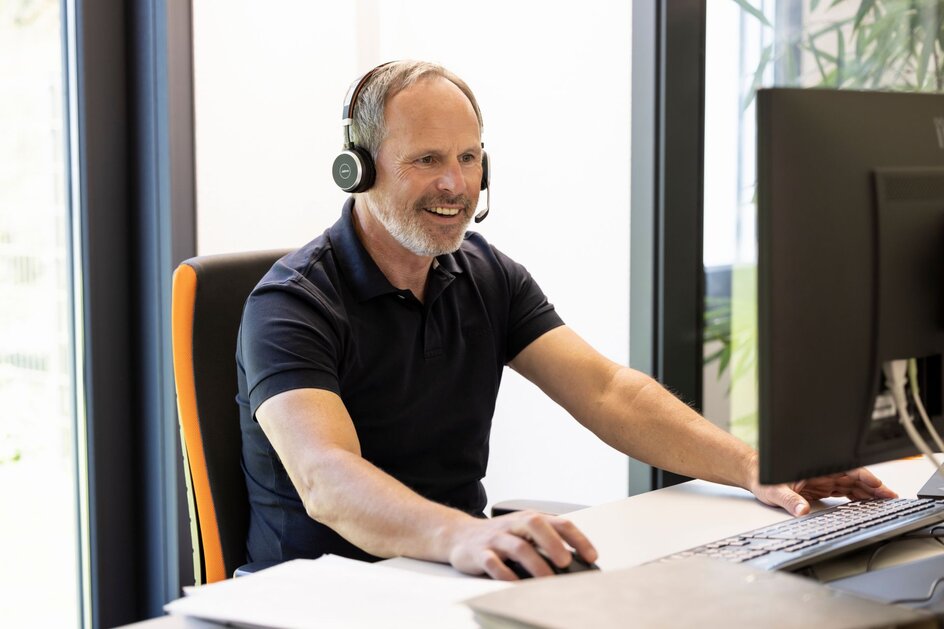 Ein Mann sitzt an einem Schreibtisch vor einem Computerbildschirm und trägt ein Headset. Er arbeitet konzentriert und lächelt, während er mit Maus und Tastatur interagiert.