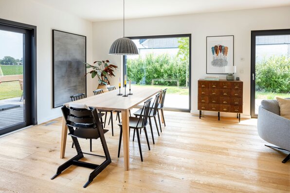 Moderner Essbereich in einem FingerHaus mit großem Holztisch, schwarzen Stühlen, heller Einrichtung und Blick in den Garten durch bodentiefe Fenster.