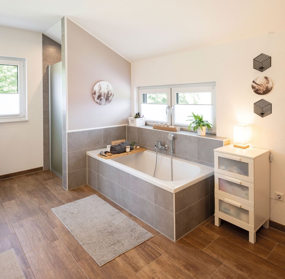 Modernes Badezimmer mit Badewanne, Glasduschwand und Holzoptikboden. Fenster, helle Fliesen und ein weißes Regal sorgen für eine freundliche Atmosphäre.