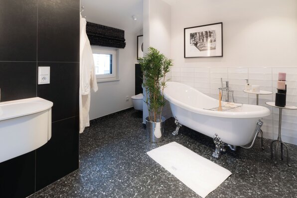 Stilvolles Badezimmer mit freistehender Badewanne auf schwarzen Fliesen im Musterhaus SENTO Bad Vilbel. Weiße Wandfliesen, Pflanzen, kleiner Beistelltisch und gerahmte Wandbilder sorgen für ein elegantes Ambiente.