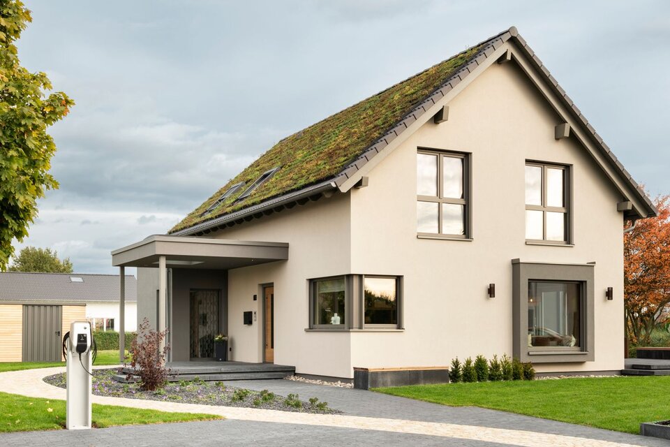 Straßenansicht des Musterhauses JUNO 300 in Mülheim-Kärlich mit Gründach, Carport-Eingang und moderner Einfahrt mit E-Ladesäule.
