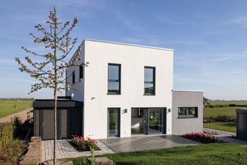 Außenansicht des FingerHaus-Musterhauses TALO in Fulda mit kubischer Architektur, Flachdach, weißer Fassade, großen bodentiefen Fenstern, Terrasse, Rasenfläche, Gartenbeeten, Baum und freistehendem Geräteschuppen unter blauem Himmel.