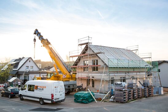 Baustelle eines zweigeschossigen Hauses mit Satteldach in Montagephase; das Gebäude ist von Gerüsten umgeben, Dachflächen sind mit Unterkonstruktion versehen. Ein großer gelber Kran steht davor, daneben ein weißer Transporter mit FingerHaus-Logo. Weitere Baumaterialien, Paletten und Werkzeuge liegen im Vordergrund, Wohnhäuser im Hintergrund.