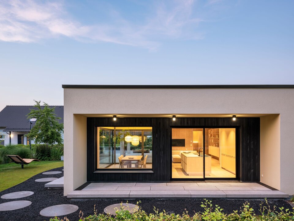 Gartenansicht des Musterhauses NIVO in Frankenberg mit überdachter Terrasse, bodentiefen Fensterfronten und beleuchtetem Wohn- und Essbereich in der Abenddämmerung.