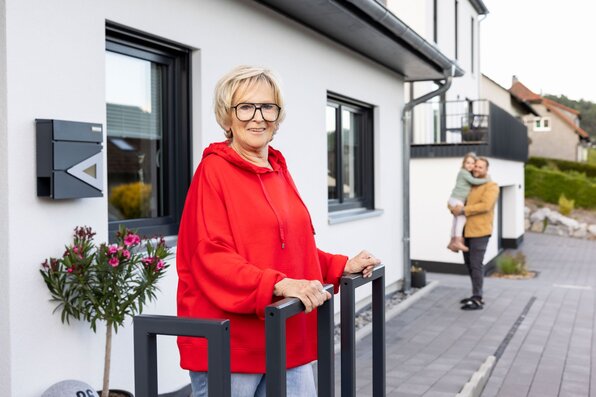 Seniorin steht am Eingangsbereich des Kundenhauses NIVO 80, während im Hintergrund ein Mann ein Kind auf dem Arm hält.