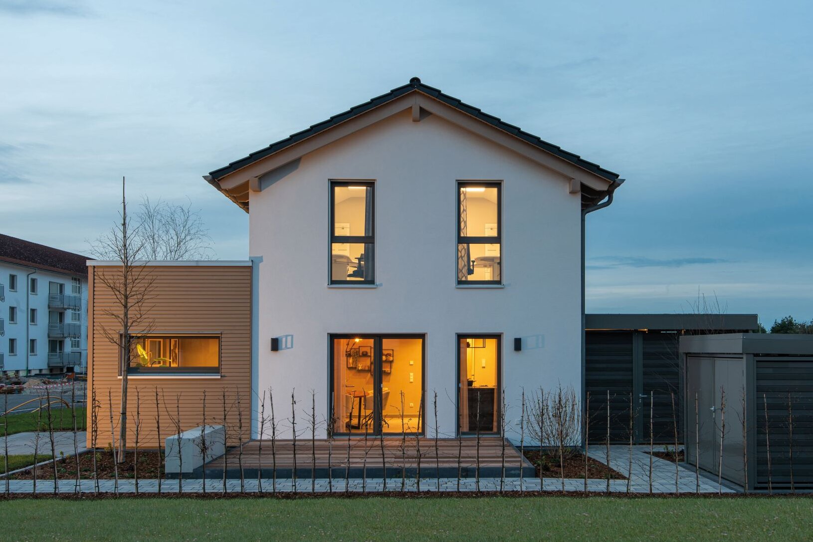 Musterhaus TALO in Babenhausen in der Abenddämmerung, warm beleuchtete Fenster zur Terrasse und moderne Anbauten mit Garage im Hintergrund.