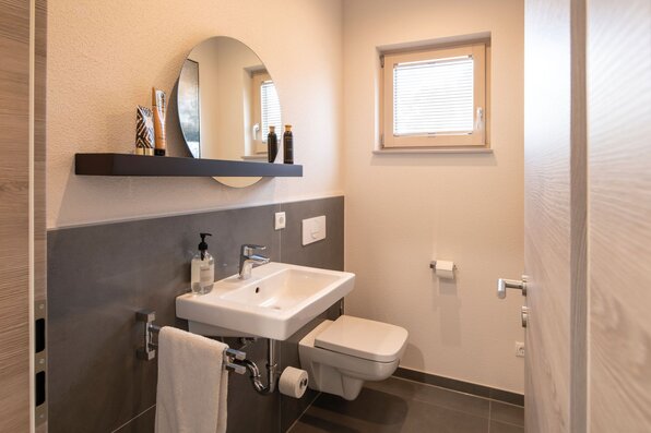 Modernes Gäste-WC im Musterhaus MEDLEY 300 B in Frankenberg mit Wand-WC, Waschbecken und rundem Spiegel.