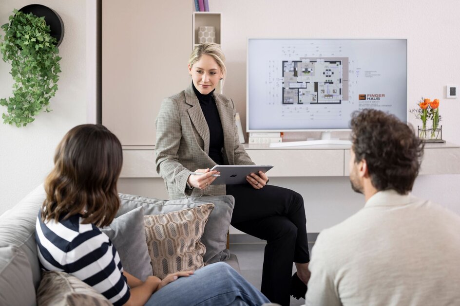 Beraterin mit Tablet erklärt Paar auf Sofa Grundriss eines FingerHaus Fertighauses auf Bildschirm im Hintergrund