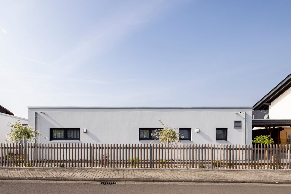 Seitenansicht eines modernen eingeschossigen Flachdachhauses mit weißer Fassade, schmalen Fenstern und Holzzaun entlang der Straße