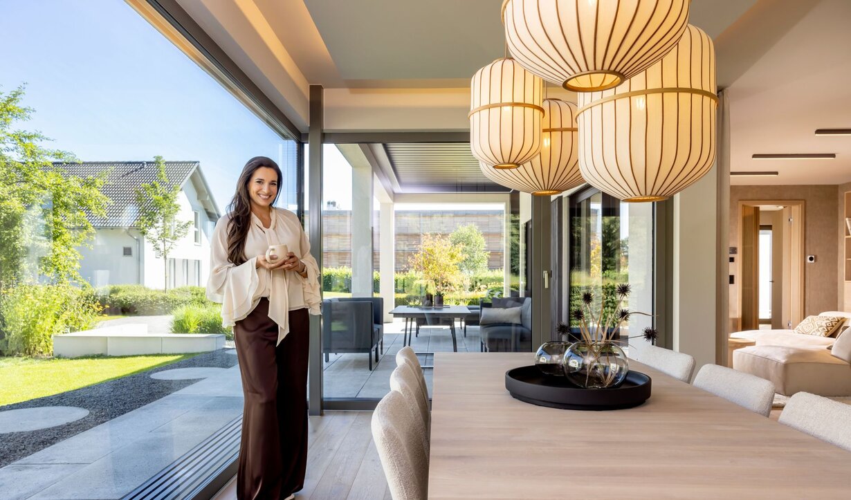 Food-Bloggerin Sally Özcan im Essbereich des Musterhauses NIVO »Sally Edition« im Musterhauspark Frankenberg mit großem Esstisch, Pendelleuchten und Blick auf die überdachte Terrasse.