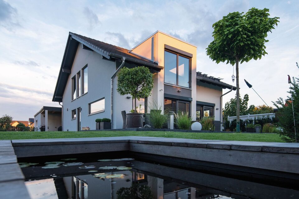 Modernes Einfamilienhaus bei Dämmerung mit großer Terrasse, gepflegtem Garten und Wasserbecken im Vordergrund.  