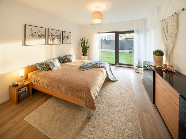 Schlafzimmer mit Doppelbett, Kommode, Teppich und großer Fensterfront mit Blick in den Garten.