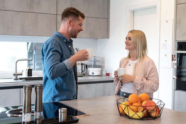 Ein Paar steht in der modernen Küche eines FingerHaus NIVO 130 und unterhält sich bei einer Tasse Kaffee. Im Vordergrund liegen Obst in einer Schale und ein Kochfeld.