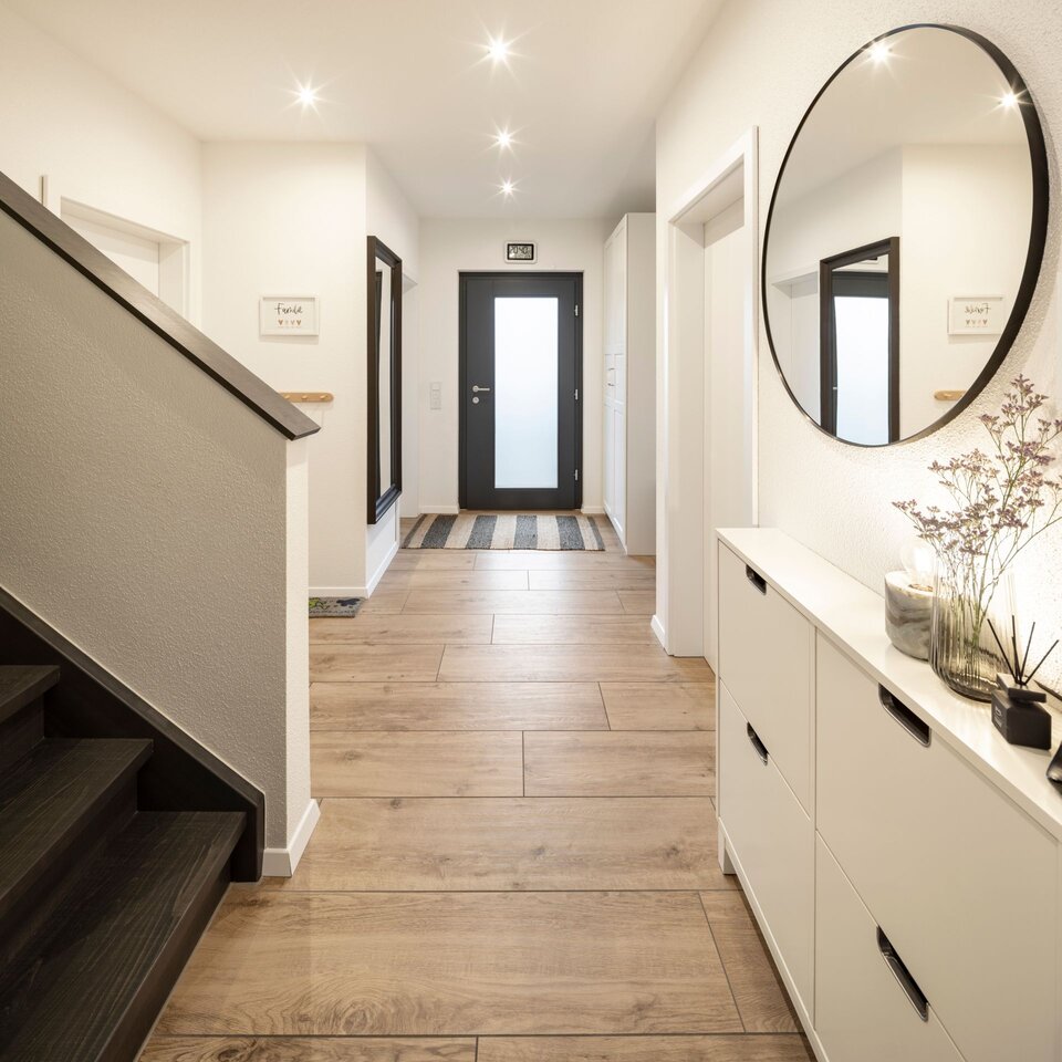 Heller Eingangsbereich mit dunkler Holztreppe, Kommode und rundem Spiegel. Flur mit grauem Boden und weißen Wänden, modern beleuchtet.
