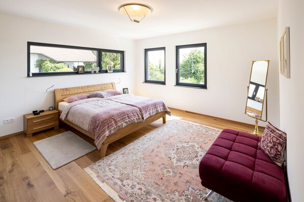 Schlafzimmer in einem frei geplanten FingerHaus mit Doppelbett aus Holz, großen Fenstern und hellem Holzboden. Ergänzt durch einen Teppich, eine Sitzbank in Bordeauxrot und einen Standspiegel.