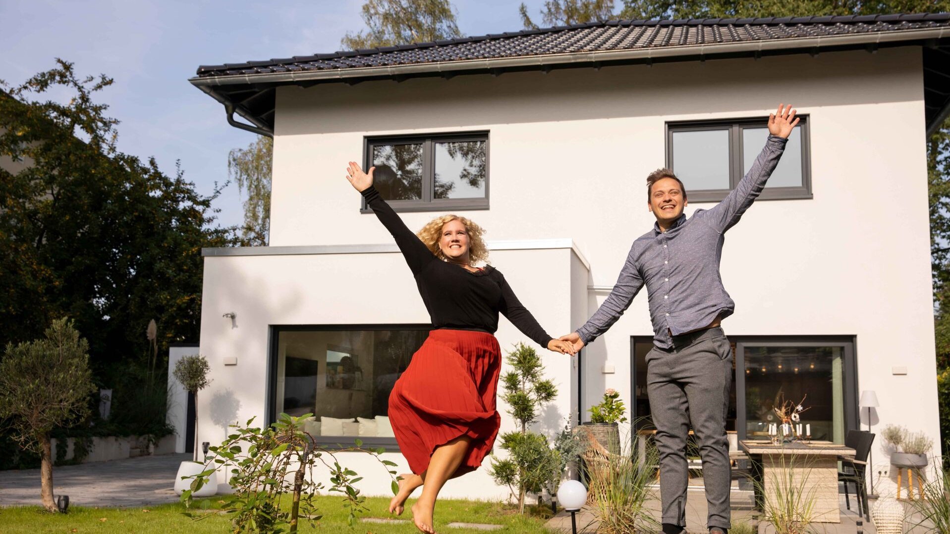 Ein Paar springt fröhlich vor seinem FingerHaus Fertighaus MEDLEY mit heller Fassade, dunklem Dach und Garten – Freude über das neue Zuhause.