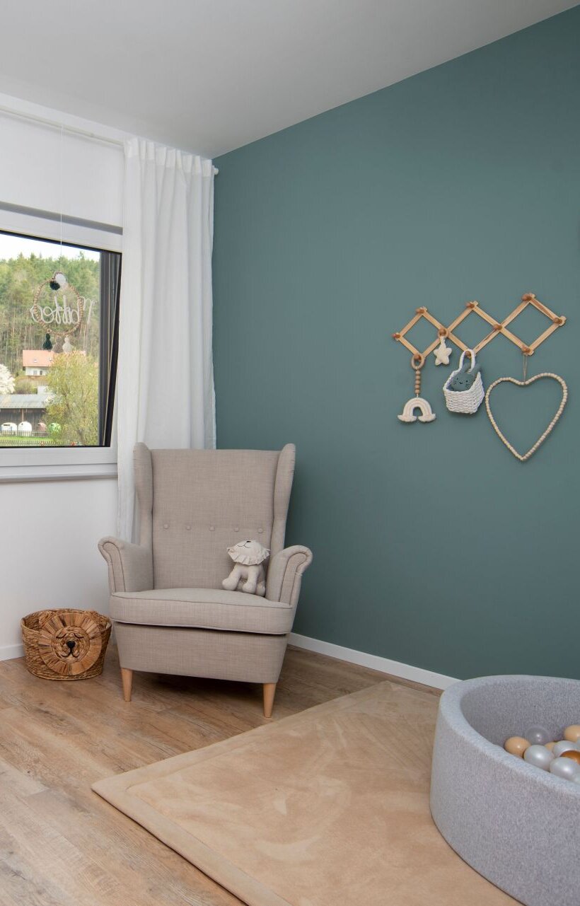 Helles Kinderzimmer in einem FingerHaus NIVO 130 mit Holz-Babybett, gemütlichem Sessel, Spielteppich, Bällchenbad und dekorativer türkisfarbener Wand.