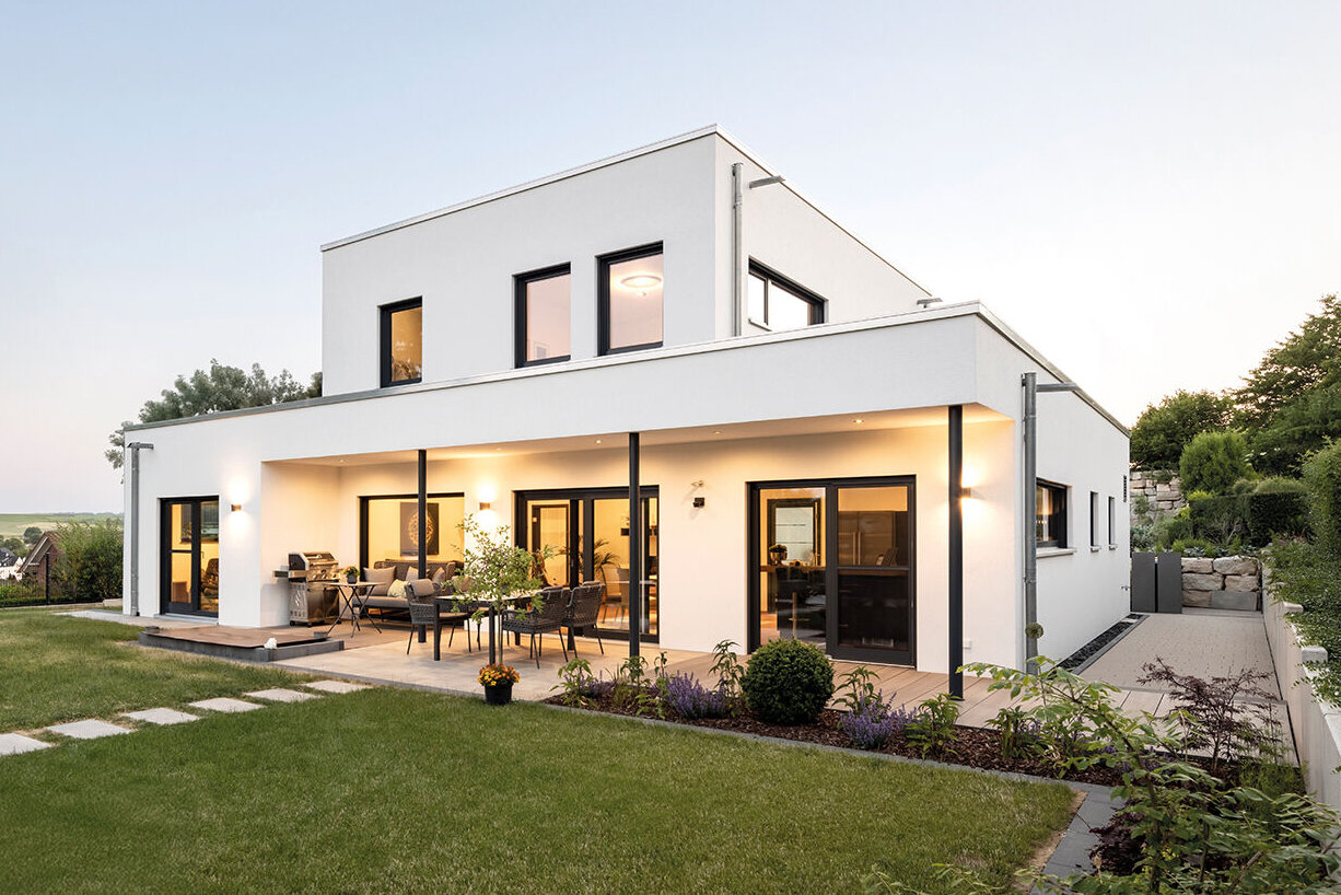 Modernes, zweigeschossiges Flachdachhaus in weißer Putzfassade, mit großen dunklen Fensterflächen, überdachter Terrasse, gepflegtem Garten mit Rasenfläche, Blumenbeeten und Hecken, aufgenommen bei Abendlicht.