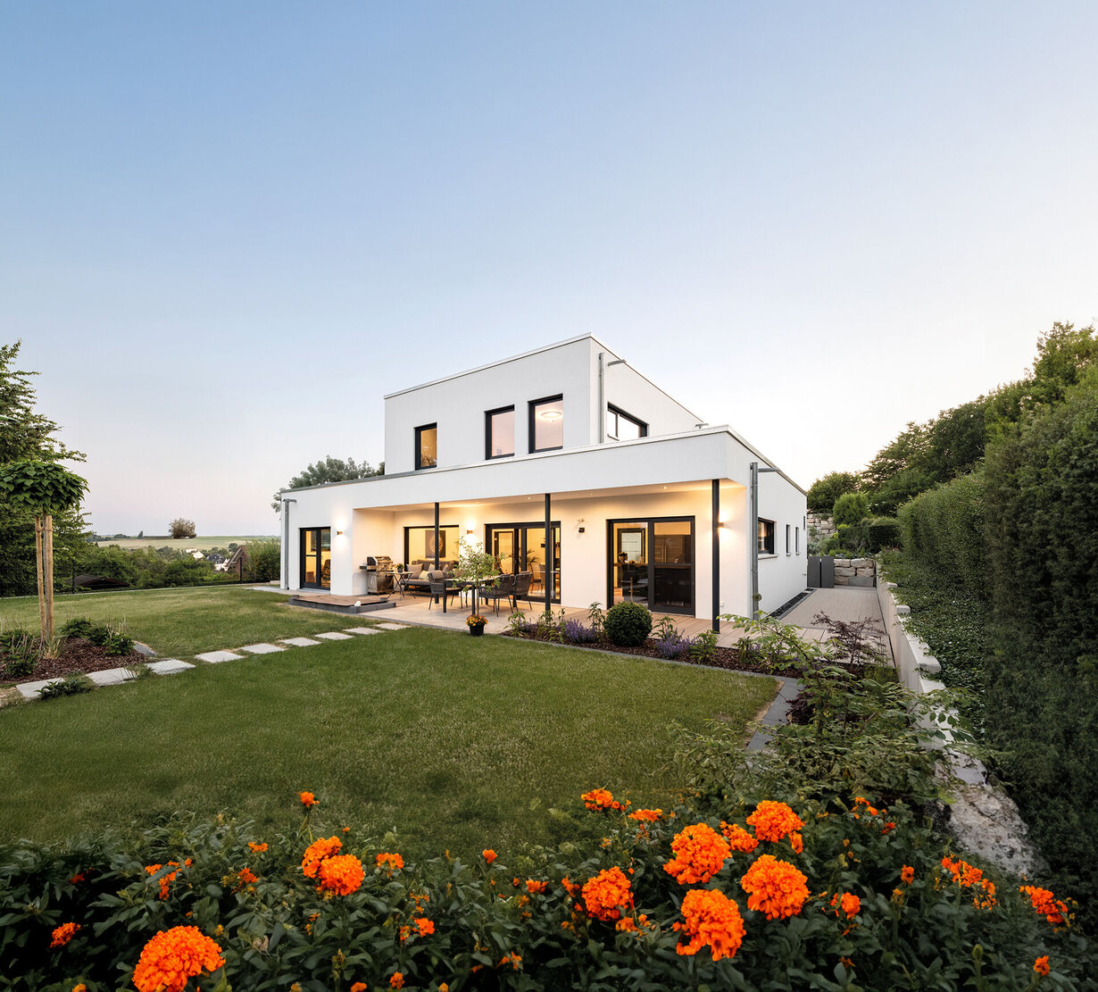 Modernes, zweigeschossiges Flachdachhaus in weißer Putzfassade, mit großen dunklen Fensterflächen, überdachter Terrasse, gepflegtem Garten mit Rasenfläche, Blumenbeeten und Hecken, aufgenommen bei Abendlicht.