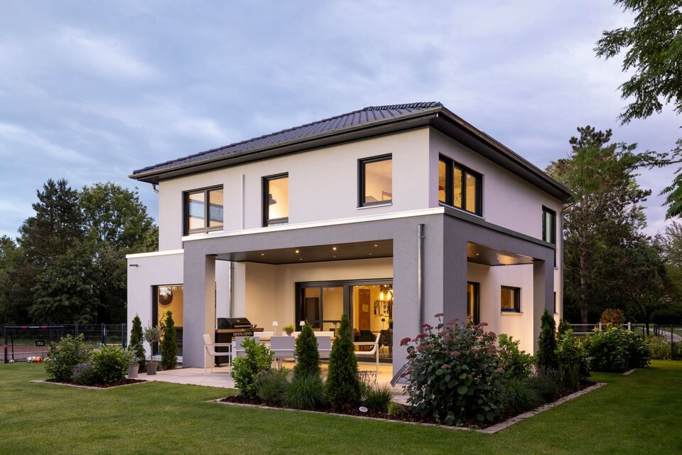 Modernes, zweigeschossiges Einfamilienhaus von FingerHaus bei Dämmerung mit heller Putzfassade und grauem Fassadenrahmen, überdachter Terrasse mit Sitzmöbeln, großen Fensterflächen, beleuchtetem Innenraum und gepflegtem Garten.