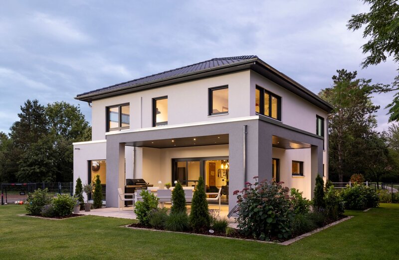 Modernes, zweigeschossiges Einfamilienhaus von FingerHaus bei Dämmerung mit heller Putzfassade und grauem Fassadenrahmen, überdachter Terrasse mit Sitzmöbeln, großen Fensterflächen, beleuchtetem Innenraum und gepflegtem Garten.