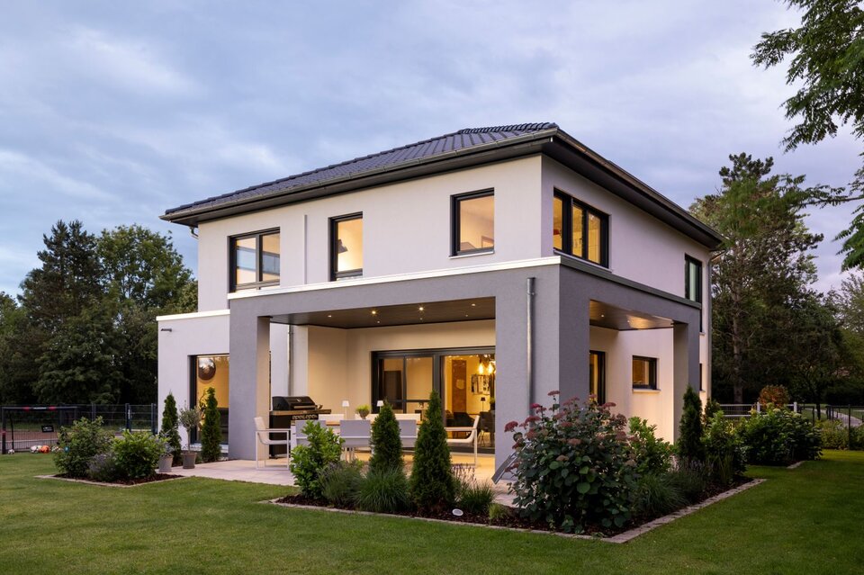 Modernes, zweigeschossiges Einfamilienhaus von FingerHaus bei Dämmerung mit heller Putzfassade und grauem Fassadenrahmen, überdachter Terrasse mit Sitzmöbeln, großen Fensterflächen, beleuchtetem Innenraum und gepflegtem Garten.