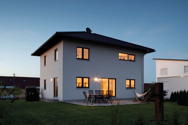 Zweigeschossiges weißes Einfamilienhaus mit Walmdach, beleuchteten Fenstern und Terrasse bei Dämmerung.