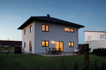 Zweigeschossiges weißes Einfamilienhaus mit Walmdach, beleuchteten Fenstern und Terrasse bei Dämmerung.