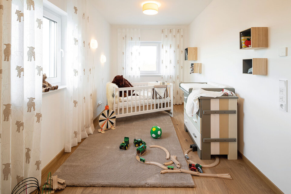 Kinderzimmer mit weißem Babybett, hellem Vorhang, Spielteppich und verschiedenen Holzspielzeugen auf dem Boden.