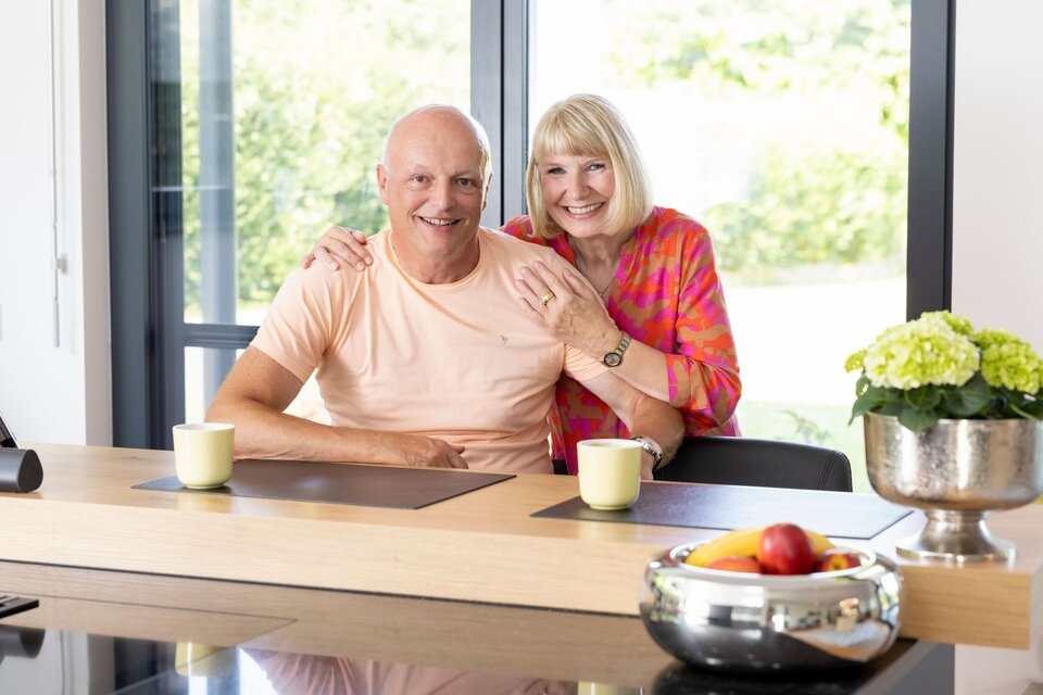 Ein Paar sitzt lächelnd am Küchentresen in einem frei geplanten FingerHaus. Auf dem Tisch stehen Kaffeetassen, eine Obstschale und eine Blumendekoration.
